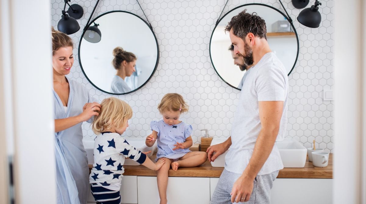 Young family in a bright bathroom—parents helping two toddlers at the sink; round mirrors on a hex-tile wall
